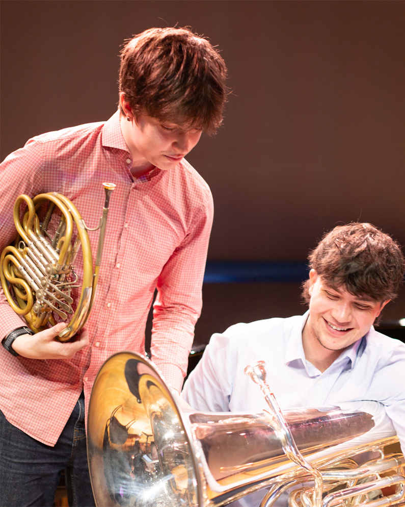 Two male students, wearing shirts, holding their brass students, looking at a mu